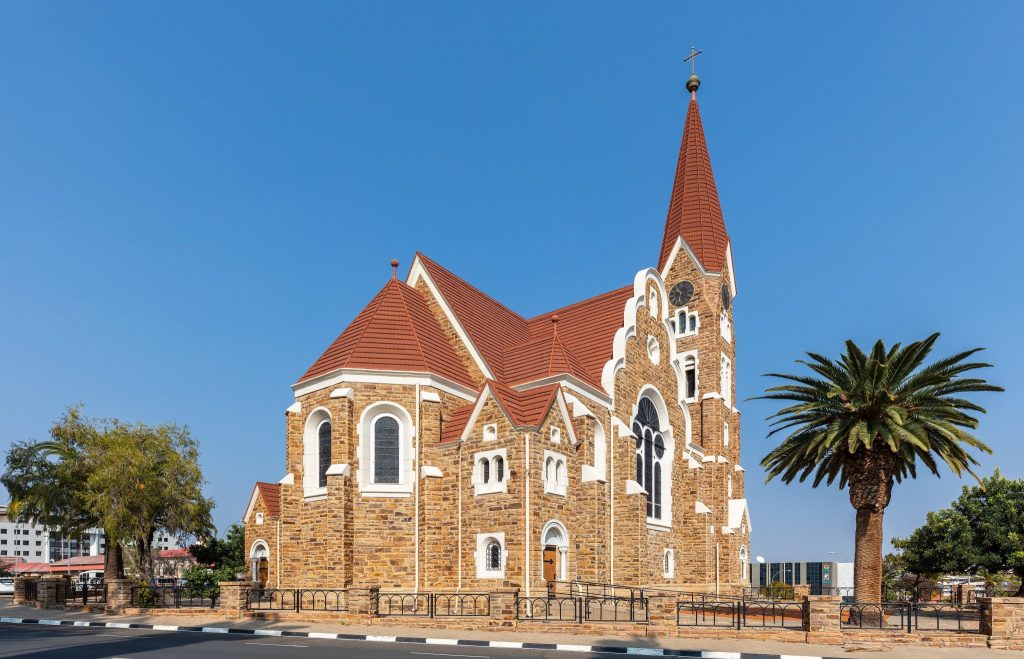 The sandstone Christuskirche in Windhoek, a Lutheran church with a red tiled roof and tall spire, beside a palm tree under a clear blue sky
