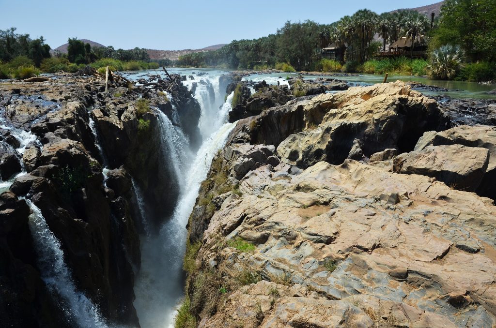A white curtain of water at Epupa Falls dropping into a narrow rock gorge on the Kunene River, makalani palms lining the far bank