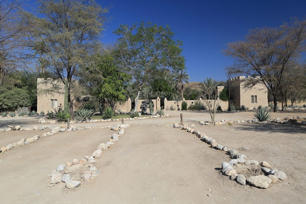 The restored mud-brick fort at Sesfontein with crenellated towers and a stone-lined gravel drive, framed by acacias and date palms in the northern Kunene
