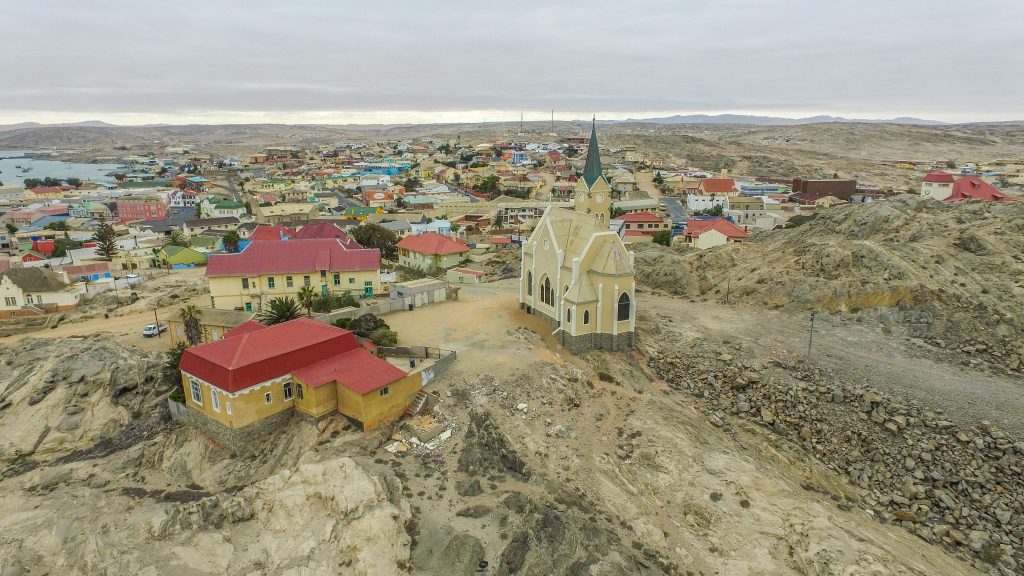 Aerial view over Lüderitz showing the Felsenkirche on its rocky outcrop above brightly painted colonial houses, with the harbour and desert hills beyond