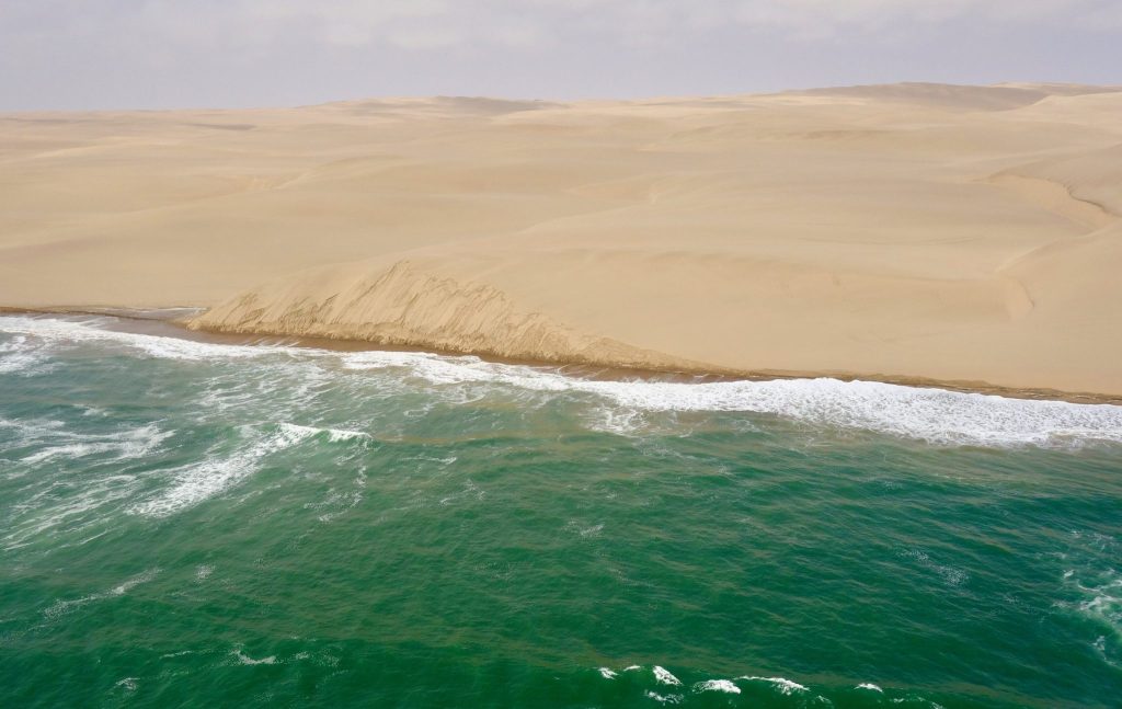 Aerial view of the Namibian coast where pale Namib dunes drop in a sand cliff straight into green Atlantic breakers along an empty shoreline