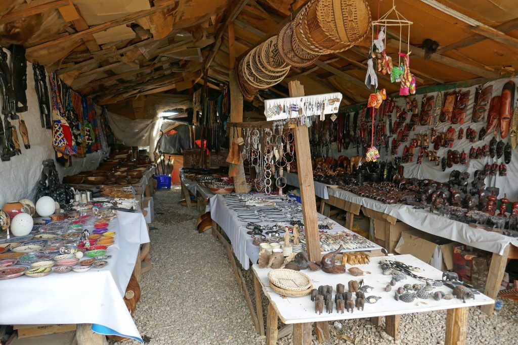 Trestle tables and wall displays at the Okahandja craft market packed with carved wooden animals, painted bowls, masks and beadwork under a rough timber roof
