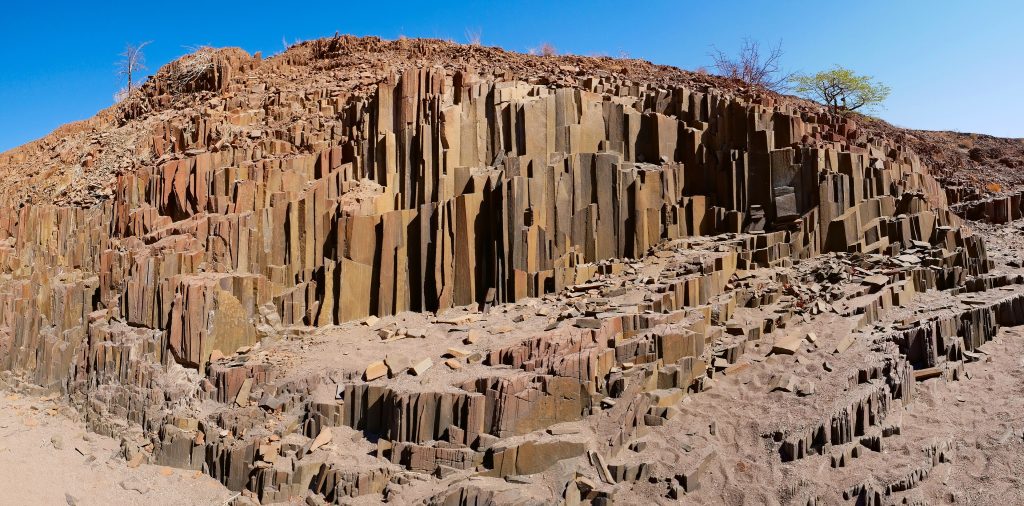 A wall of tightly packed vertical basalt columns, the Organ Pipes formation in Damaraland, with a green tree on the ridge above