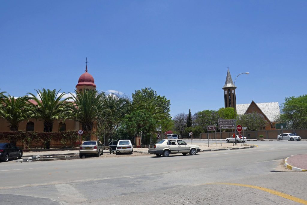A quiet street corner in Otjiwarongo with parked cars, a red-domed church tower among palm trees on the left and a tall stone church spire on the right