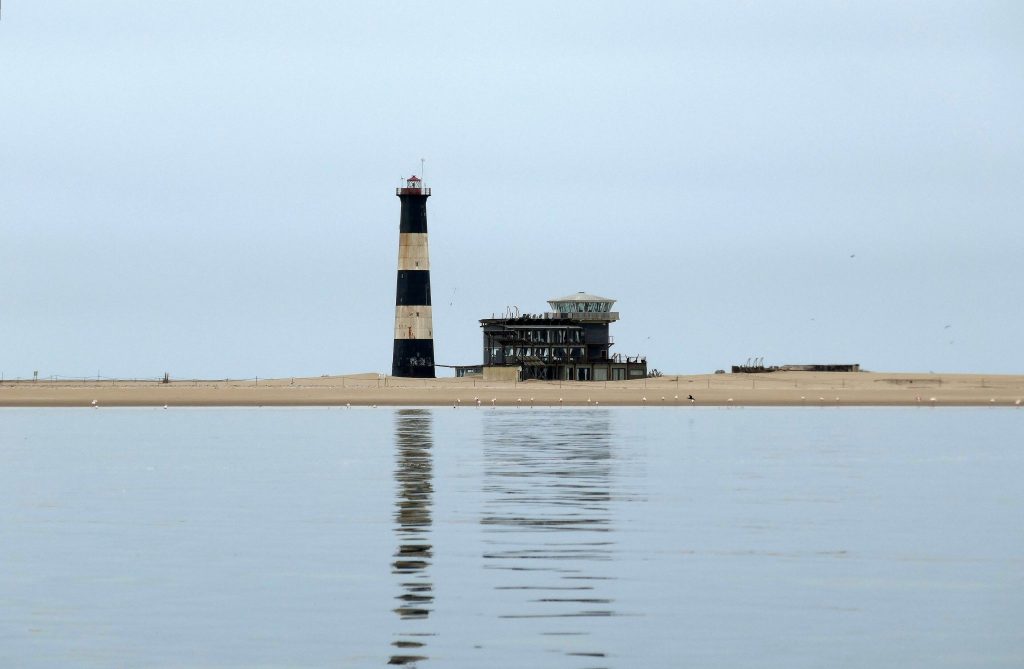 The black and white banded Pelican Point lighthouse and lodge on a low sandspit across the still water of Walvis Bay lagoon, with flamingos feeding along the shoreline