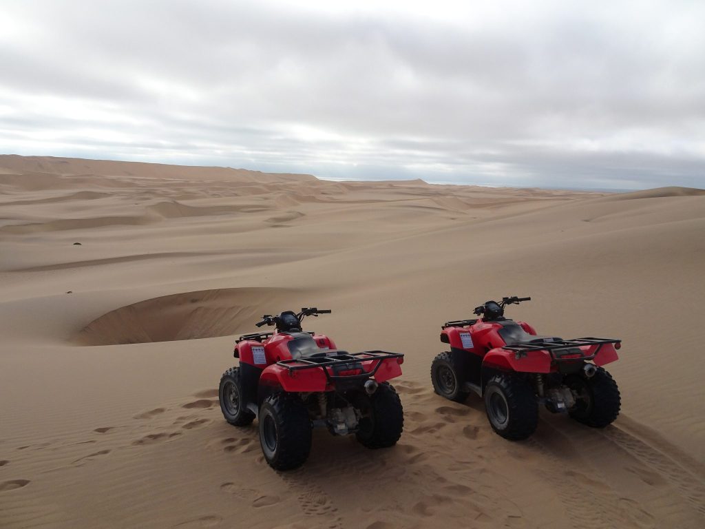 Two red quad bikes parked on a sand dune in the desert outside Swakopmund, with rolling dunes stretching to the horizon under an overcast sky