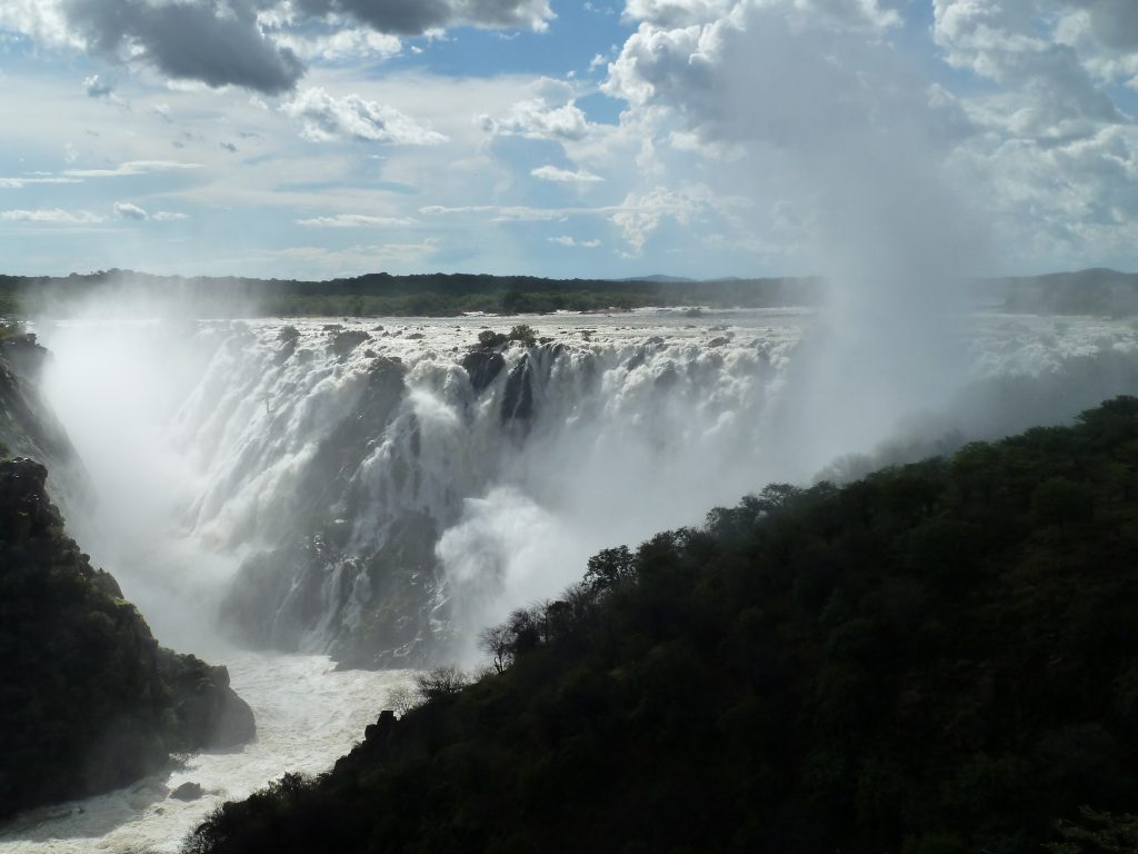 Ruacana Falls in full flood, a broad wall of white water dropping into a green gorge beneath a tall cloud of spray