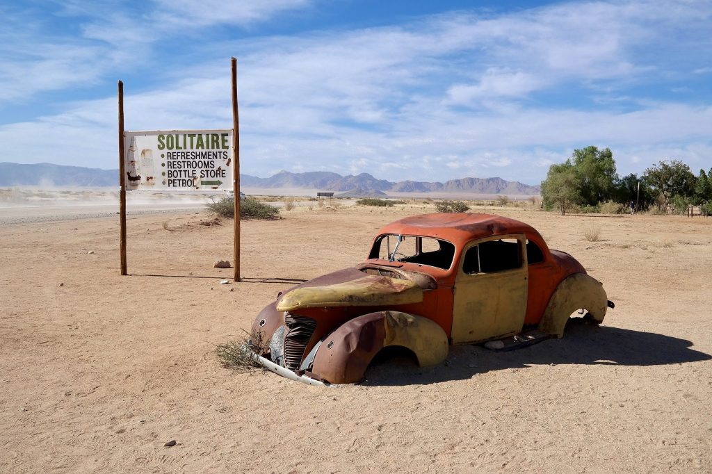 A rusting 1940s car body half sunk in the sand beside the painted Solitaire signboard advertising refreshments and petrol, with desert mountains behind