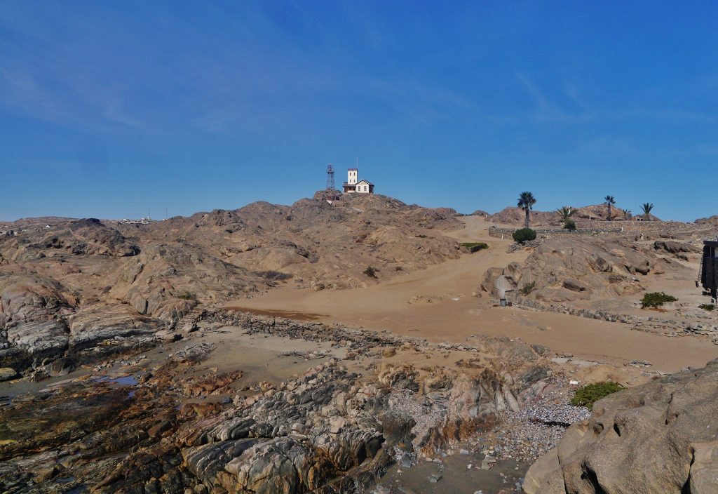 The rocky peninsula of Shark Island at Lüderitz, with a white signal station building on the granite ridge above a sandy track and tidal rock pools