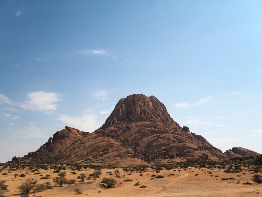 The bare granite dome of Spitzkoppe rising abruptly from a flat scrub-dotted desert plain under a pale blue sky