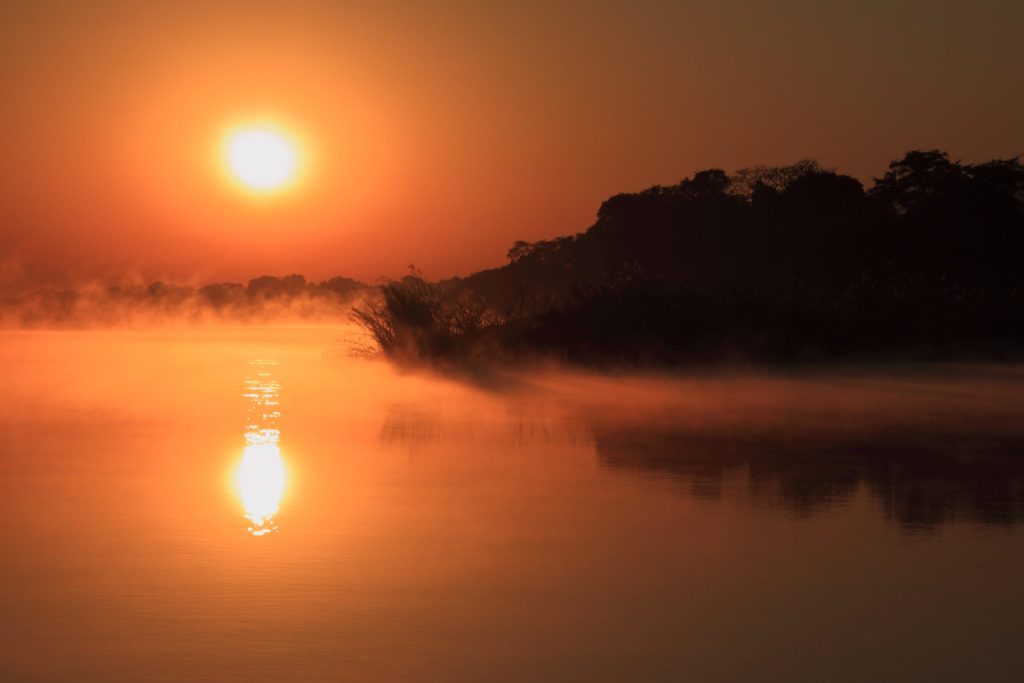 The sun rising through orange haze over the Zambezi River in the Caprivi Strip, with mist drifting off the water and a dark tree-lined bank in silhouette