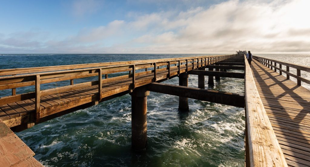 The wooden jetty at Swakopmund running out over breaking Atlantic surf, lit by low sun with a single walker in the distance