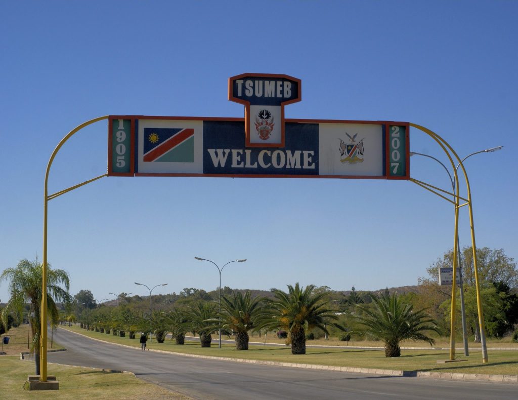 A yellow steel arch carrying a Tsumeb welcome sign with the Namibian flag and town coat of arms, spanning a palm-lined avenue into the town