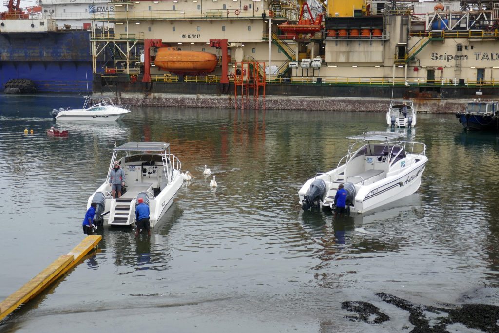 White marine tour boats moored in Walvis Bay harbour with white pelicans swimming between them and a large offshore drilling vessel at the quay behind