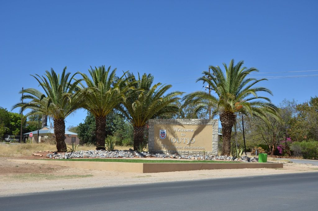 A stone-framed Welcome to Outjo sign set in a landscaped bed of rocks and aloes beneath tall date palms at the edge of the town road